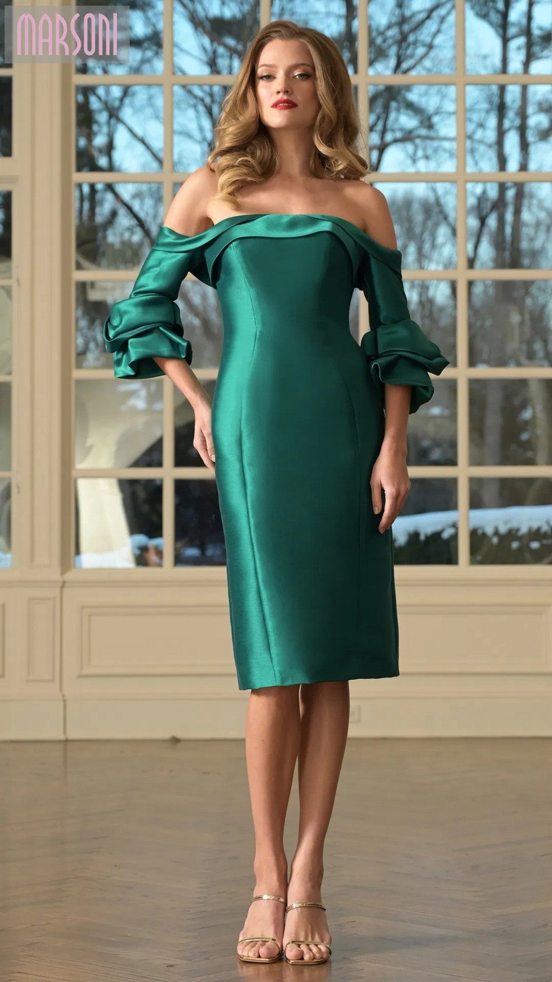 Woman wearing the Marsoni MV1370, a knee-length semi-formal emerald dress with off-the-shoulder ruffled sleeves and sheath silhouette, standing indoors by large windows.